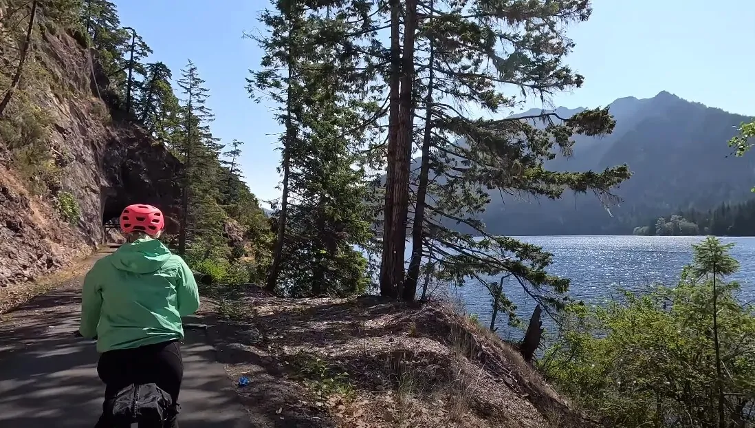A paved trail winds through a lush forest, leading to a tranquil lake