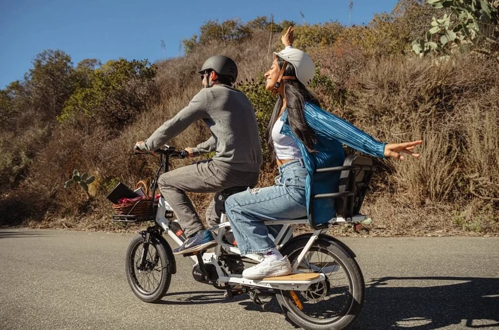 Two people riding a cargo e-bike on a sunny countryside road, both wearing helmets and enjoying the ride