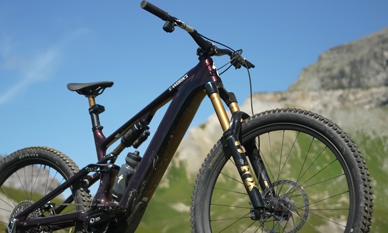 Dark purple S-Works full-suspension mountain bike against a bright blue sky and rocky mountain backdrop