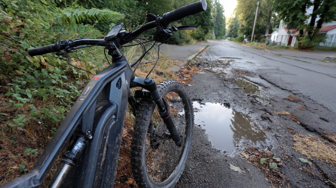 An electric bike stopped on a cracked roadside with puddles and trees nearby