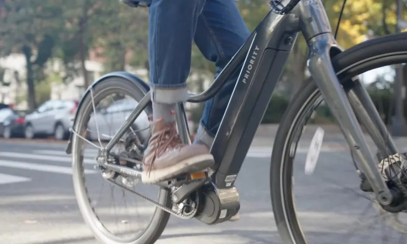 A person wearing jeans and brown sneakers rides a black Priority e-bike on a street