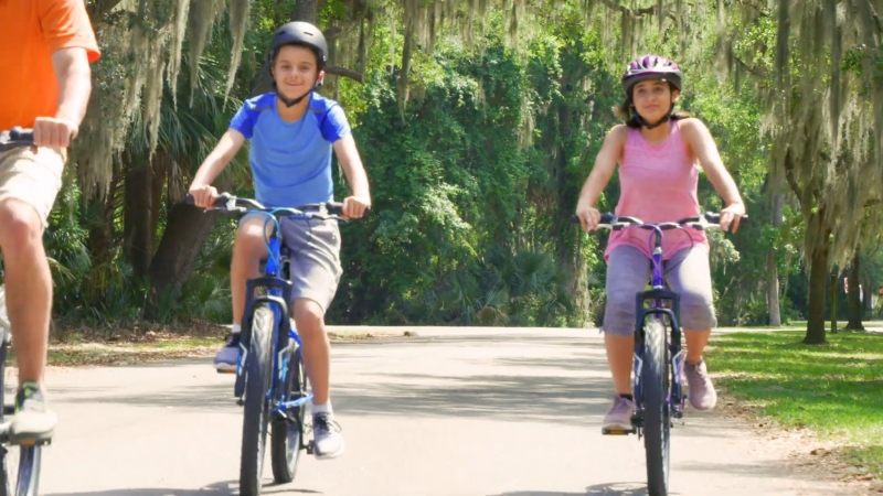 Two kids wearing helmets riding electric bikes on a paved park trail