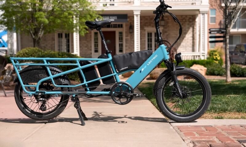 A blue electric cargo bike with a rear storage compartment stands on a sunny sidewalk