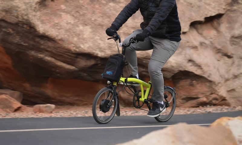 A person rides a bright green folding bike along a smooth road with large reddish rocks in the background