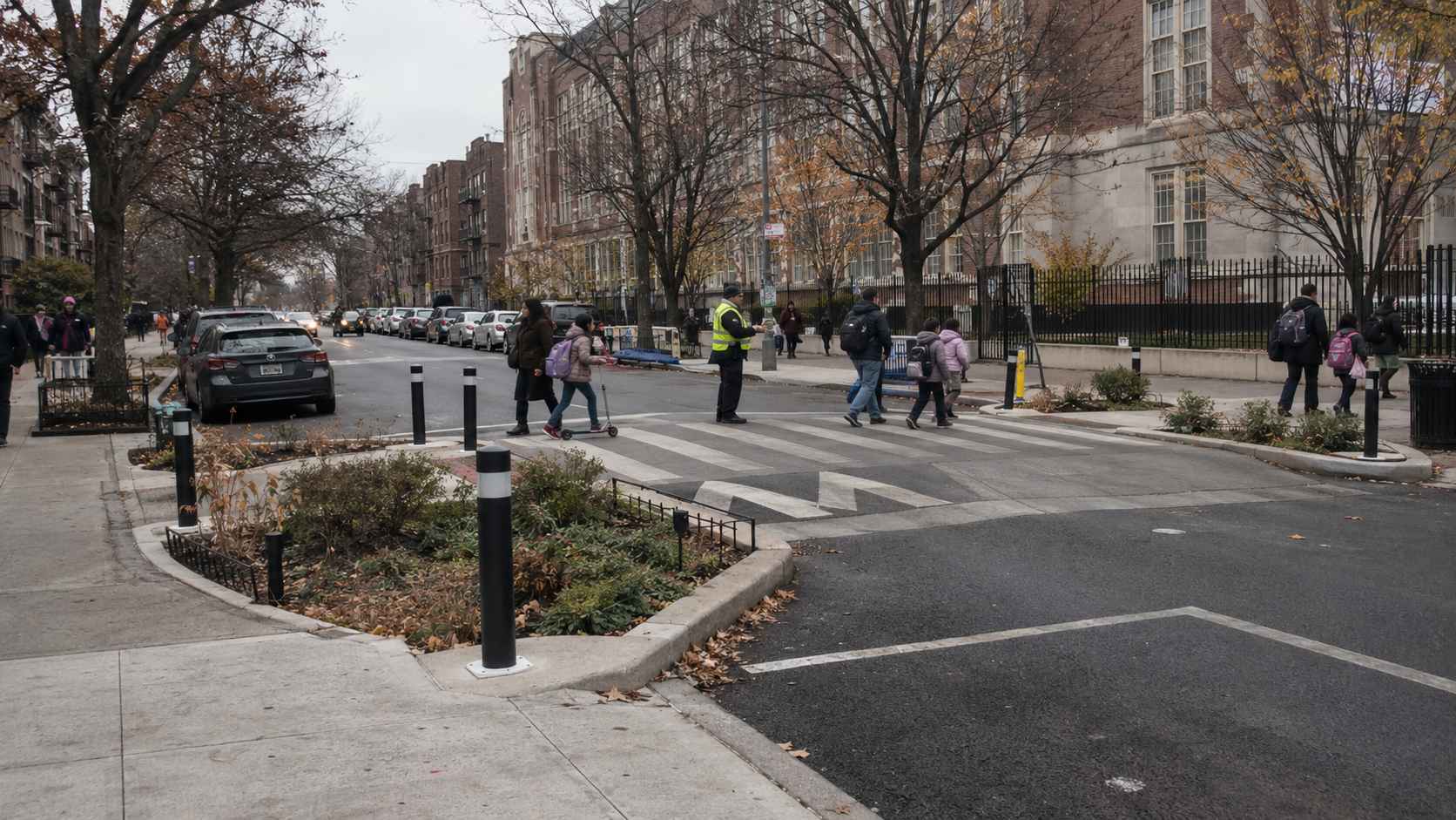 Students cross a wide pedestrian crossing near a school as a crossing guard watches.